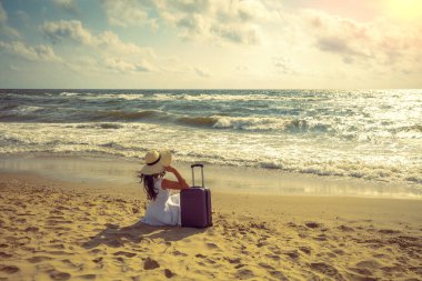 Hasır şapkalı ve seyahat valizli genç mutlu kadın yazın sahilde oturur. Kadın hasır şapkayı tutuyor ve denize bakıyor. Seyahat, iyi tatiller, yaz tatili konsepti