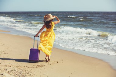 Uzun elbiseli ve şapkalı, seyahat valizli mutlu bir kadın yazın sahilde yürüyor. Kadın denize bakmaktadır. Seyahat ve iyi tatiller.;