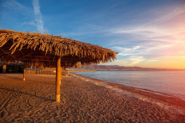 Güneş şemsiyeli tropik plaj manzarası. Gün batımında plajda saman güneş şemsiyeleri. Eilat, İsrail