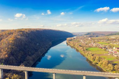 Güneşli bir sonbahar gününde nehrin üzerindeki bir yol köprüsünün havadan görünüşü. Nehir kıyısındaki köy.