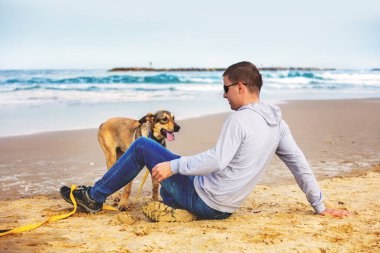 Genç bir adam köpeğiyle sahilde oturuyor.