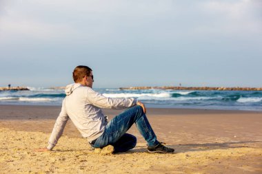 Genç bir adam sahilde oturur ve denize bakar.