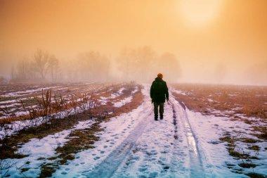Bir adam sisli bir kış sabahı kırsalda toprak bir yol boyunca yürür. Arazi ilk karla kaplı.