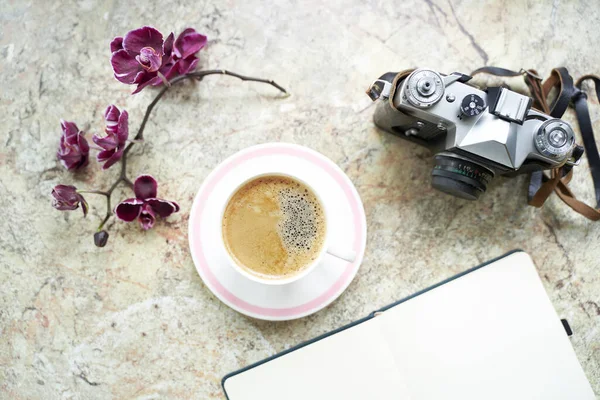 Taza de café con orquídea, cámara analógica retro, bloc de notas plano sobre mesa de mármol — Foto de Stock