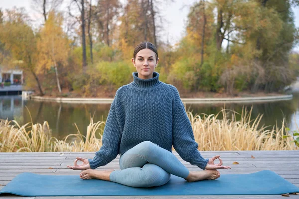 Young pretty woman doing yoga poses in the park autumn next to the lake shore