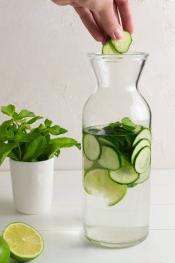 Bir el, salatalık, nane filizi ve detoks içeceği ile doldurulmuş bir sürahi taze suya birkaç parça salatalık atar. Sağlık, spor, sağlıklı gıda konsepti. Dikey yönelim.