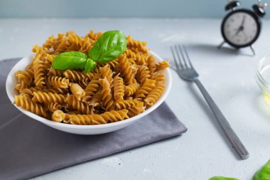 Gri arka planda saati olan gri bir peçetenin üzerindeki beyaz bir kasede çiğ fusilli. Makarna yeme zamanı. Seçici odaklanma. Yatay oryantasyon. Tam tahıllı makarna.