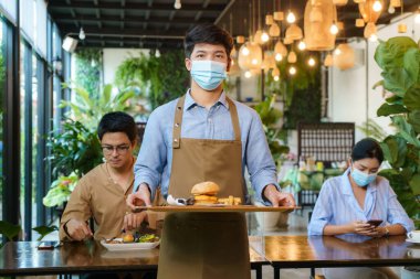 Portre çekici Asyalı garson yüz maskesi takıyor ve arka plandaki müşteriye yemek servisi için yüz kalkanı tutuyor. Yeni normal restoran konsepti