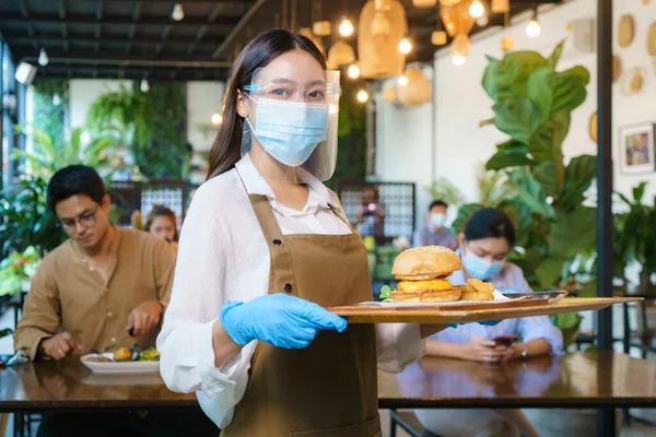 Portrait attractive asian waitress wear face mask and face shield holding food tray to serving meal to customer with custome in background. New normal restaurant concept