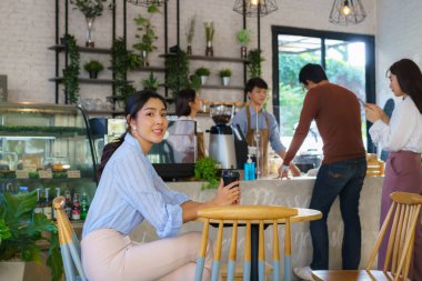 Kafede oturan Asyalı kadın kahve içerken kendini mutlu hissediyor ve diğer insanlar da baristayla içecek siparişi veriyor. 