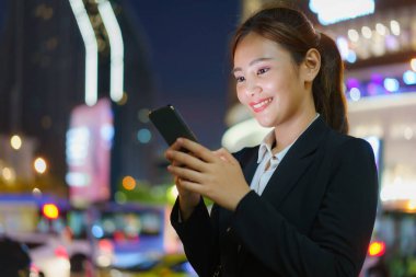 Bangkok, Tayland 'da geceleri arka planda ofis binaları olan caddede cep telefonu kullanan Asyalı yönetici kadın.