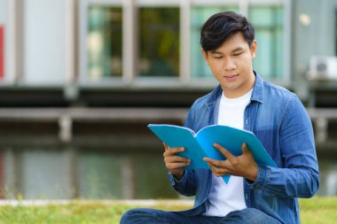 Asyalı üniversite öğrencisinin portresi kampüste çimlerin üzerinde mutlu görünüyor ve parkta kitap okuyor.