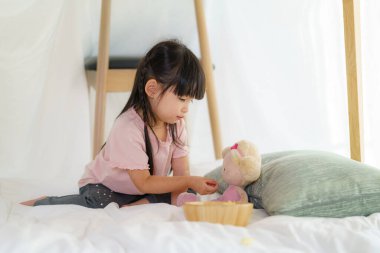 Asyalı şirin küçük kız, diğer aile üyelerinden saklanmak ve yaratıcı bir şekilde oynamaları için evdeki battaniye odasında otururken oyuncak bebeğini oynar ve abur cubur yer..
