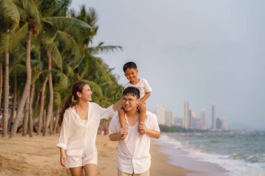 Babası oğlunu sırtında taşıyan Asyalı bir aile ve annesi yazın Tayland 'da tatildeyken deniz kenarında hindistan cevizi ağaçlarıyla birlikte yürüyorlar.