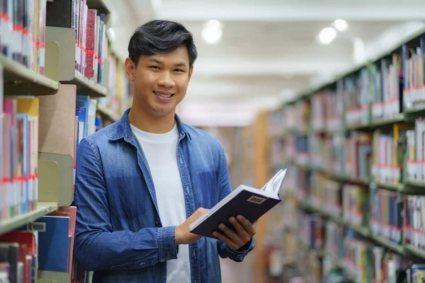 Portrait of Smart Asian man university student reading book and looking at camera between bookshelves in campus library with copyspace