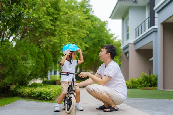 アジアの父親は息子のためにヘルメットをかぶっていて 近所の庭で自転車に乗る方法を子供に教えています — ストック写真