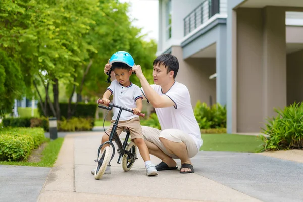 アジアの父親は息子のためにヘルメットをかぶっていて 近所の庭で自転車に乗る方法を子供に教えています — ストック写真