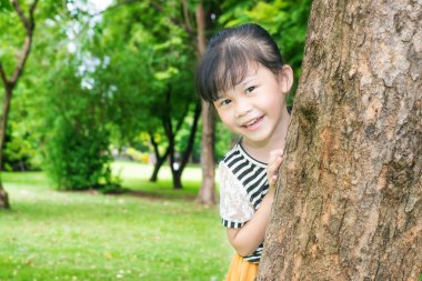 Asyalı küçük kız parkta bir ağacın arkasında gülümseyen