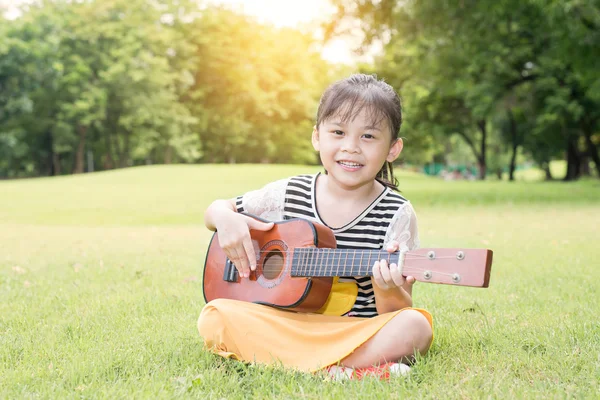 Asian dziewczynka siedzi na trawy i grać ukulele w parku — Zdjęcie stockowe