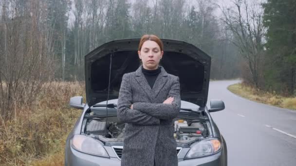 Slim woman at the background of broken car in the forest. Stressed female driver standing in front of open hood. Breakage, problem. — ストック動画