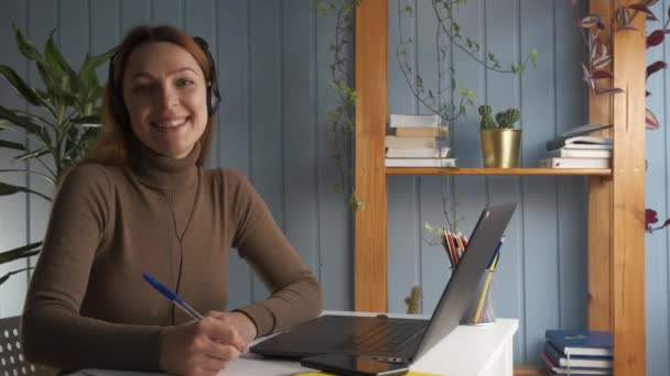 Mulher sentar-se na mesa segurar caneta escreve no copybook sorriso olhar para a câmera, inteligente excelente retrato estudante diligente, fácil e conveniente processo de auto-educação usando fone de ouvido e laptop — Vídeo de Stock