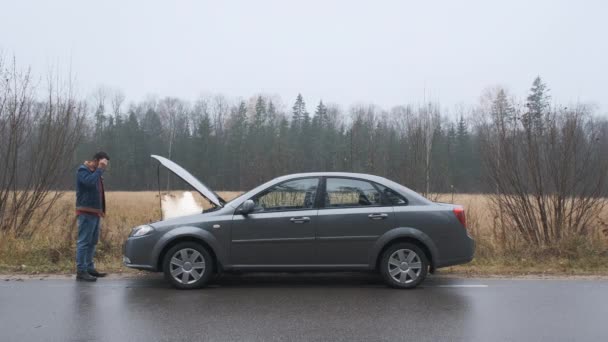 Triste homme debout sur la route près de la voiture cassée. De la fumée sort du moteur. Besoin d'aide. Service de voiture. Service de remorquage. — Video