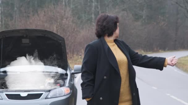 A woman stands by the road near a broken car. The woman is trying to stop someone for help. — Stock Video