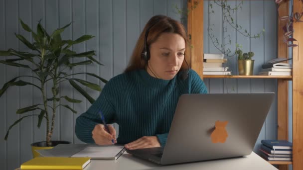 Student dziewczyna siedzieć przy biurku nosić słuchawki patrzeć na ekranie komputera odnotowując w zeszycie ćwiczeń studiuje przygotować esej używać on-line łącze internetowe, informacje wyszukiwania, koncepcja samokształcenia — Wideo stockowe