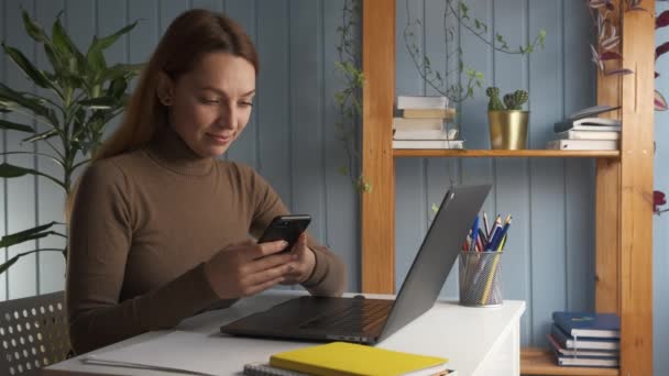 Ung kvinna sitter vid skrivbordet distraherad från studier själv utbildning verksamhet chatta med vän genom budbärare ansökan. Anställda vilar på arbetsplatsen håller smarttelefon köpa i e-handel webbplatser — Stockvideo