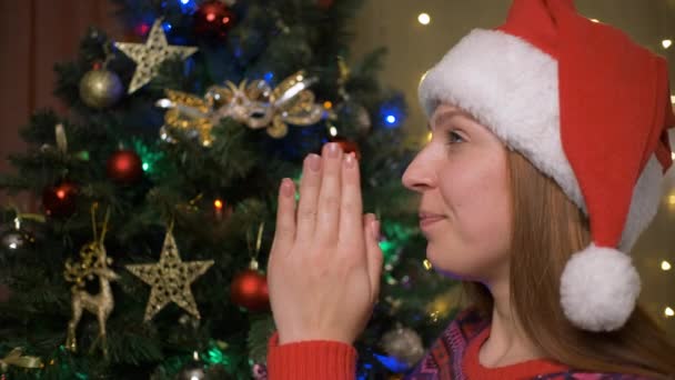 年轻的女人在圣诞树旁祈祷圣诞节礼物。戴着圣诞礼帽的女人正在梦想着圣诞节的圣诞礼物. — 图库视频影像