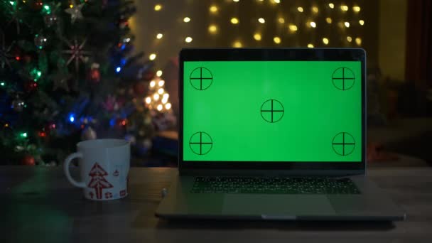 Nowoczesny laptop z kawą na drewnianym stole. Zielony ekran i choinka z ozdobami i migające światła — Wideo stockowe