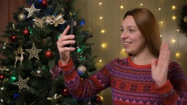 Noel arifesi video konferansı düzenleyen bir kadın uzaktan akrabalarını tebrik ediyor. Yanıp sönen ışıkların yanında oturuyor. Noel ağacı, modern teknoloji konsepti.