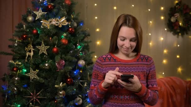 漂亮的女人接近装饰着圣诞树的圣诞树，用智能手机和朋友聊天阅读新年祝福笑起来很开心。圣诞销售电子商务用户概念 — 图库视频影像