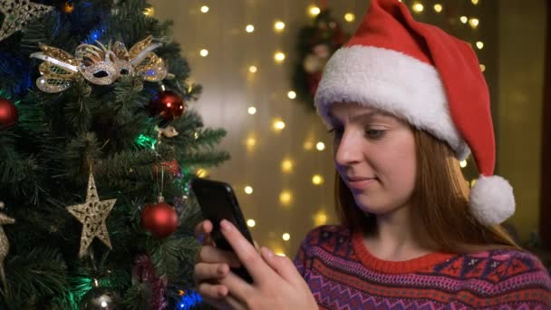 Ritratto di giovane bella donna con cappello di Babbo Natale in maglione invernale digitando messaggio sul telefono cellulare — Video Stock