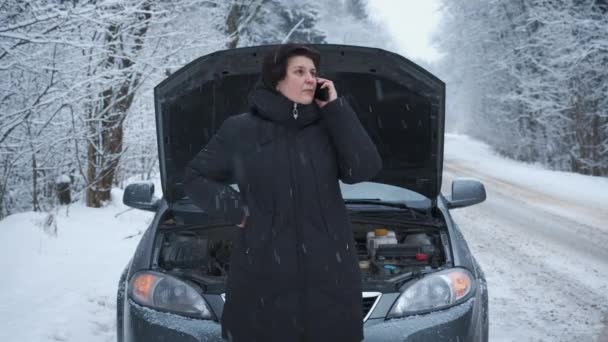 Γυναίκα στο δάσος το χειμώνα στέκεται στο δρόμο με ένα σπασμένο αυτοκίνητο, άνοιξε το καπό, μιλώντας στο τηλέφωνο, περιμένοντας για βοήθεια, ο συναγερμός του αυτοκινήτου είναι ενεργοποιημένος — Αρχείο Βίντεο