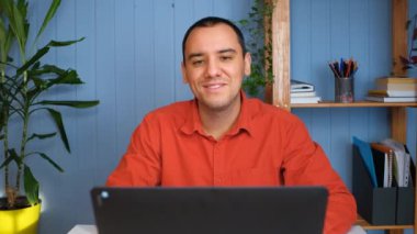 Mutlu genç iş adamının portresi, ofis masasında oturuyor ve dizüstü bilgisayarda çalışıyor. Gülümser ve kameraya bakar..