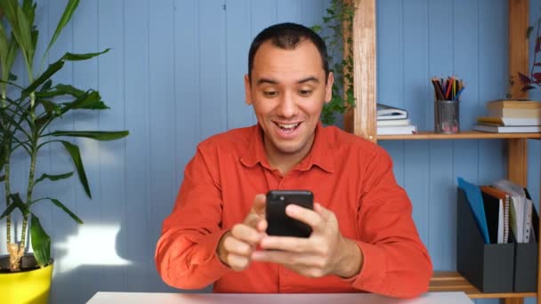 Νεαρός επιχειρηματίας παίρνει επιτυχημένα νέα, κάθεται στο τραπέζι με smartphone στο γραφείο στο σπίτι. — Αρχείο Βίντεο