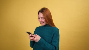 Cep telefonunda mesaj yazan, gülümseyen, sohbet eden, sosyal ağda iletişim kuran, uygulamayı kullanan mutlu genç bir kadın. sarı arkaplanda izole