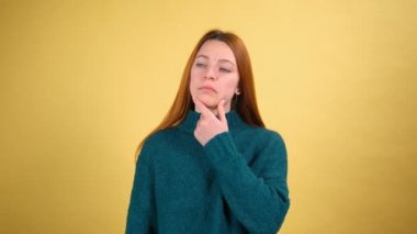 Düşünceli genç bir kadın, yeşil kazaklı, kızıl saçlı, çenesini ovuşturan ve düşünceli bir ifadeyle yan yana bakan. Çözüme kafa yormak, şüpheli bir soru. Stüdyo çekimi sarı arkaplanda izole edildi