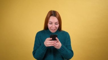 Oldukça çekici kızıl saçlı kıvırcık saçlı genç kız cep telefonuyla sohbet ederek cep telefonuna bakıyor. İyi eğlenceler. Stüdyoda sarı arka planda izole edilmiş. Halk yaşam tarzı konsepti.