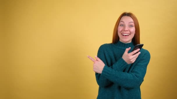 緑のセーターを着た20代の若い楽しい女性は、携帯電話の指差し指をワークスペースに置いて手をつないですごいと言います。スタジオで黄色の背景に隔離されたスペースをコピーします。人々のライフスタイルコンセプト — ストック動画