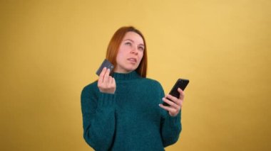 Cep telefonu kullanmaktan heyecan duyan genç bir kadın kredi kartı kullanıyor ve harika bir fikri var, evet diyor. Sarı arka plandaki stüdyoda izole edilmiş. Halk yaşam tarzı konsepti.