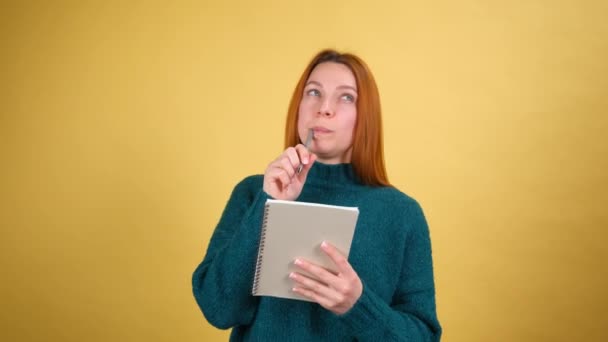 Задумчивая творческая молодая женщина в зеленом свитере обдумывает будущий бизнес-план, делает заметки в бумажном блокноте, пишет творческие идеи и улыбается вдохновляюще счастливым. в закрытом помещении — стоковое видео