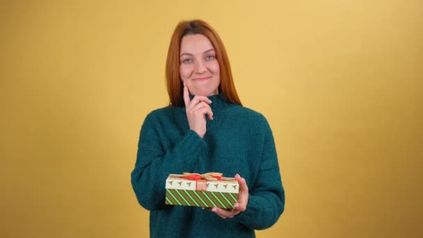 Junge Frau schüttelt verpackte Geschenkschachtel, hält das Ohr am Ohr, hört zu, was drinnen ist, schaut mit neugierig verwirrtem Gesichtsausdruck. Vereinzelt auf gelbem Hintergrund — Stockvideo