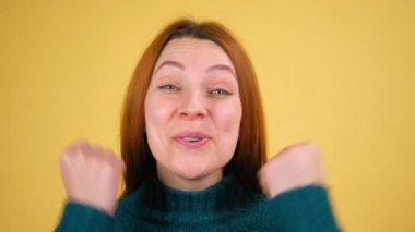 Gülümseyen, kızıl saçlı, güzel bir kadın kazanan bir jest yapıyor. Stüdyoda sarı arka planda izole edilmiş 