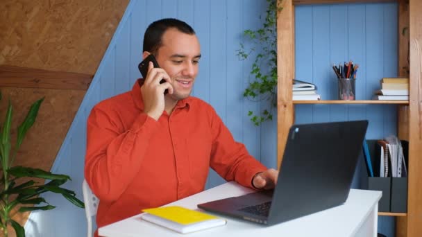 Sonriente joven hombre de negocios profesional hablando por teléfono usando el ordenador portátil sentarse en el escritorio de la oficina en casa — Vídeos de Stock