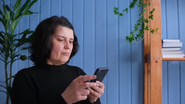 Mujer mira a la pantalla del teléfono inteligente y se siente muy contento, ganar en la lotería, recibir una buena noticia. Una dama madura triunfando en casa — Vídeos de Stock