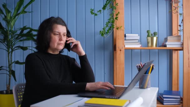 Женщина средних лет разговаривает по телефону и работает в домашнем офисе. — стоковое видео