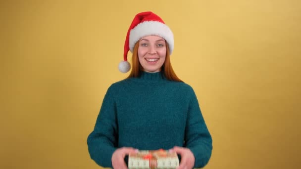 Semestertradition. Charmig ung kvinna i Santa hatt ger julklapp till kameran och ler sött, grattis på julafton, nyår. inomhus studio isolerad gul bakgrund — Stockvideo