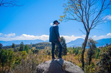 Gri kayanın üstünde duran genç adam manzarası var.